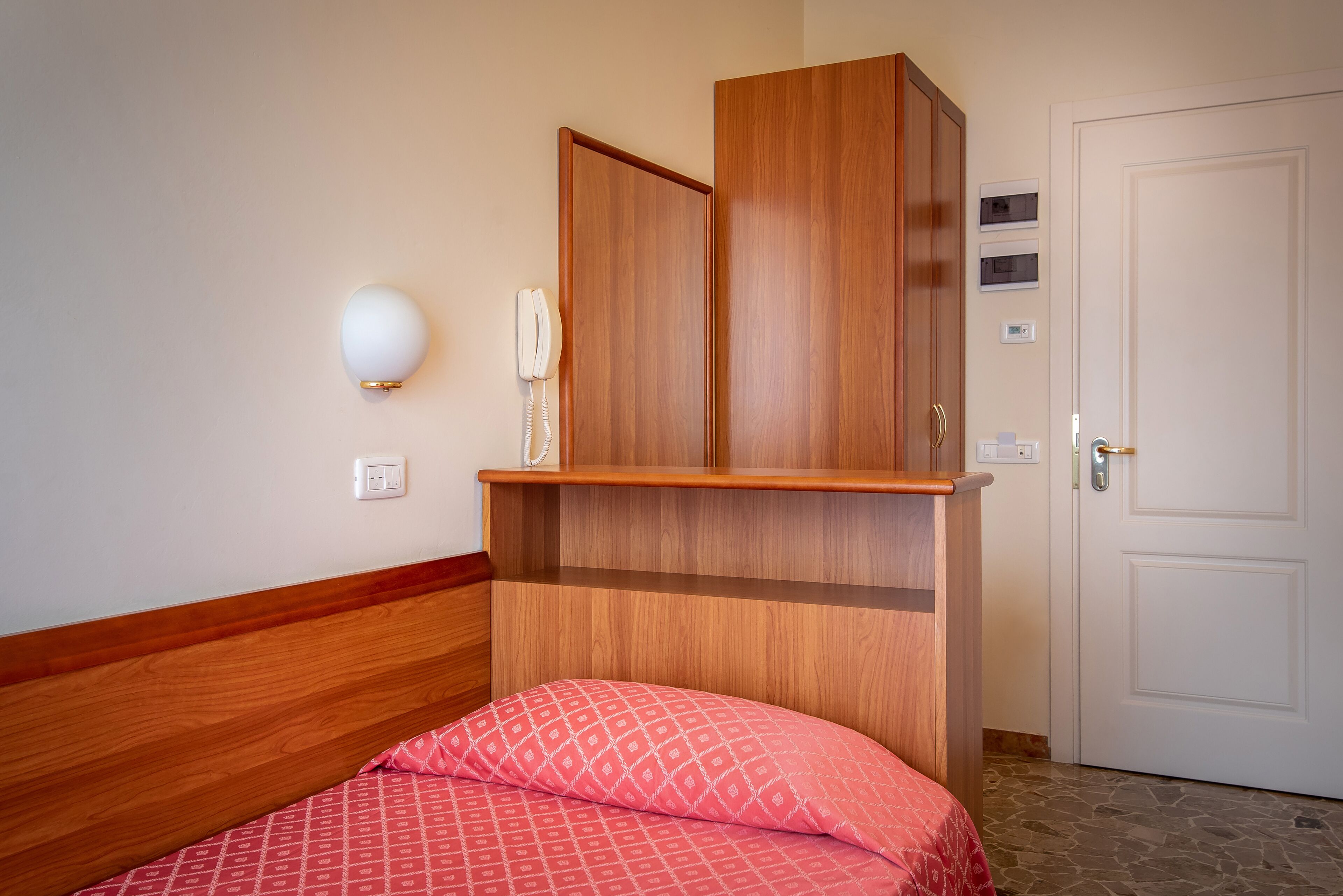 Quarto com estrutura de cama de madeira, colcha vermelha estampada, guarda-roupa de madeira, telefone de apoio e porta branca.