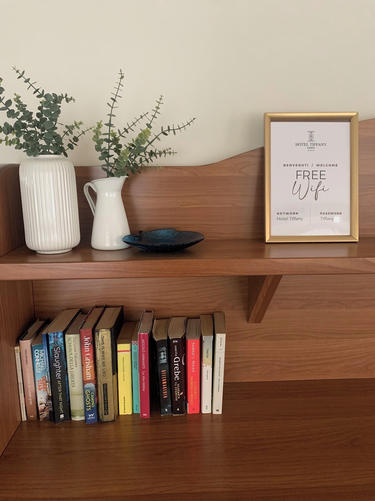 A wooden shelf with a framed sign offering free wifi, a white vase with green plants, and a row of books.
