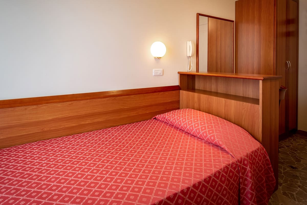 Hotellihuone, jossa on puinen sängynpääty, kuviollinen punainen peitto sängyllä, puhelin yöpöydällä ja peili puisen lipaston yläpuolella.