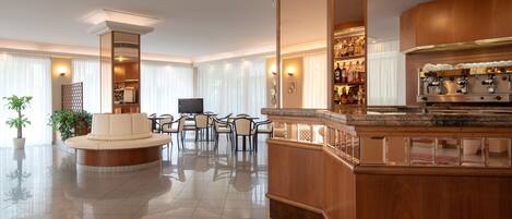 A spacious hotel lobby with a central seating area, a bar with a coffee machine, and a television.