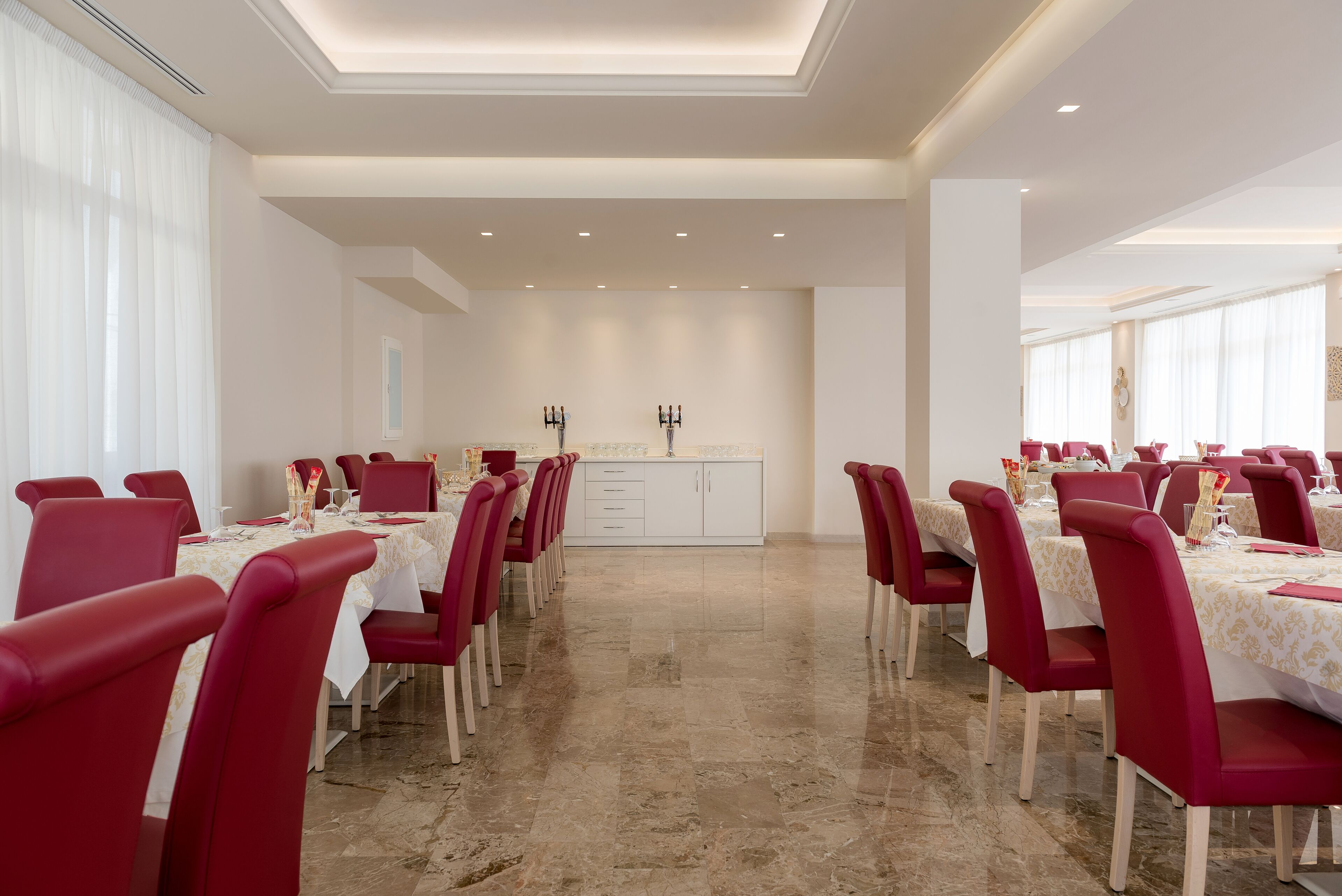 Een eethoek met rode stoelen en witte tafels, een centraal kookeiland met spoelbak, en een plafond met inbouwverlichting.
