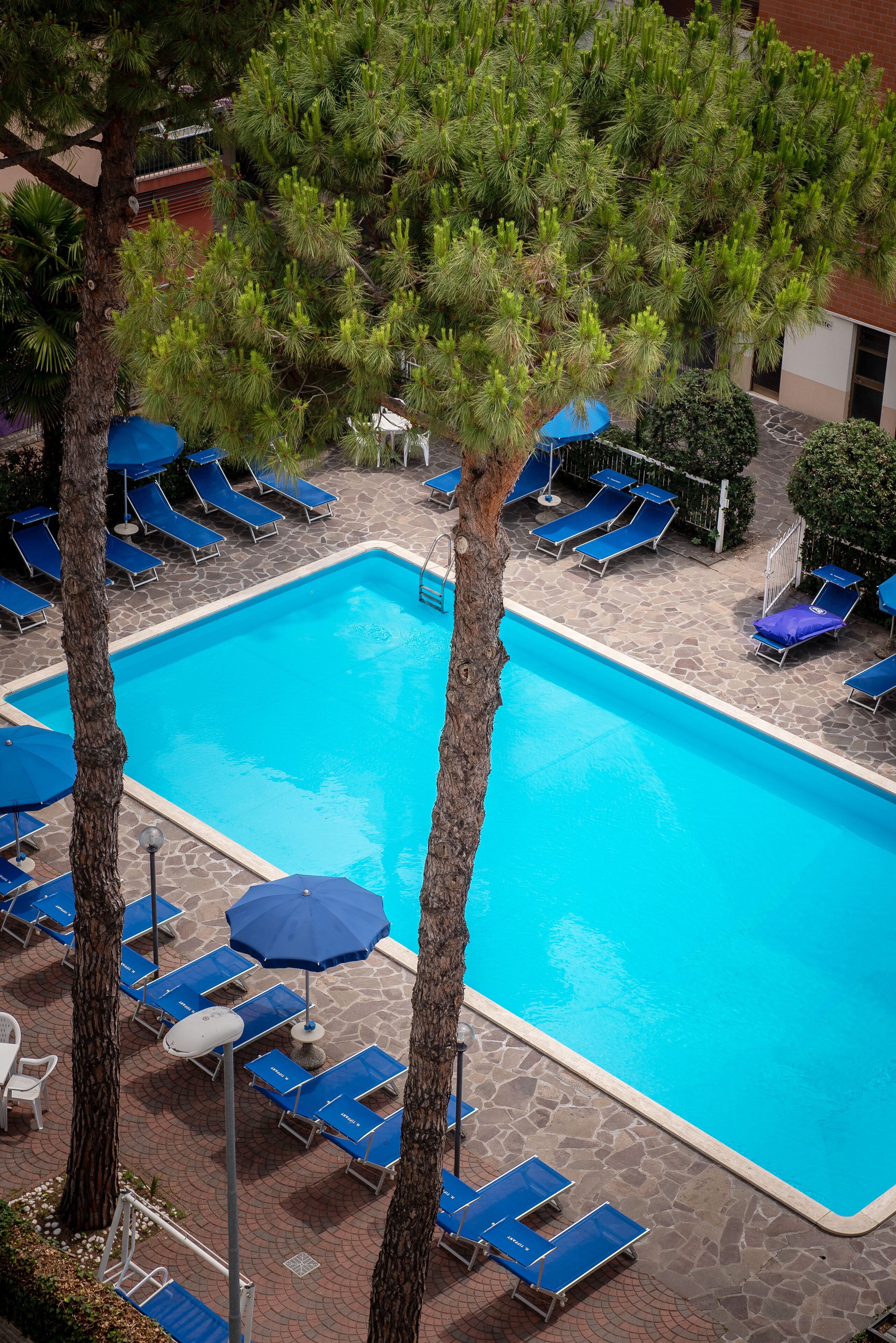 A swimming pool with blue lounge chairs and umbrellas, surrounded by trees and a stone patio.