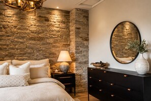 Een slaapkamer met een stenen muur, een bed met wit beddengoed, een nachtkastje met een lamp, een dressoir en een spiegel.