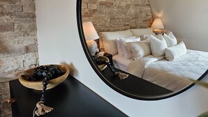 Une chambre à coucher avec un lit, un miroir rond, une table de chevet en bois sur laquelle se trouve un bol rempli d’objets noirs, et un mur en briques.