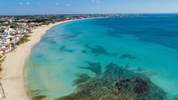 An aerial view of a coastal area with a sandy beach, clear turquoise waters, and a dense residential area with houses.
