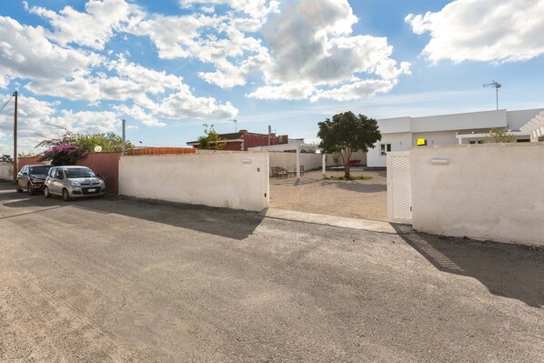 A parking area with a white wall and a gate, a car, and a building with a flat roof in the background.