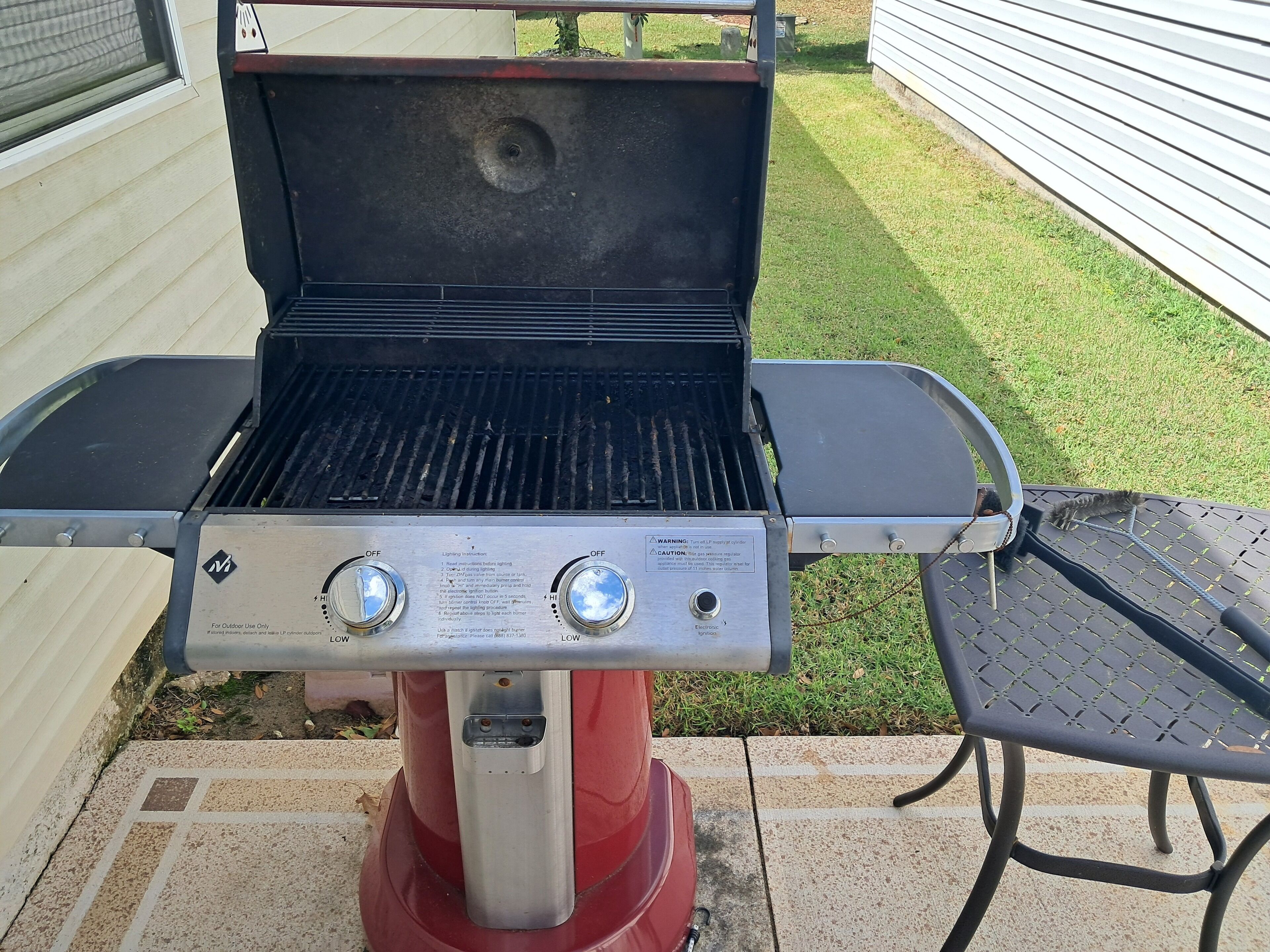 A portable grill with two control knobs and a digital display, set on a patio with a metal table and a grassy lawn in the background.