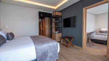 A modern bedroom with a large bed, a wooden wardrobe, a TV mounted on the wall, and a small seating area.