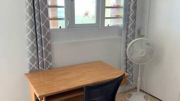 A room with a wooden desk, a black office chair, a standing fan, and a window with patterned curtains.