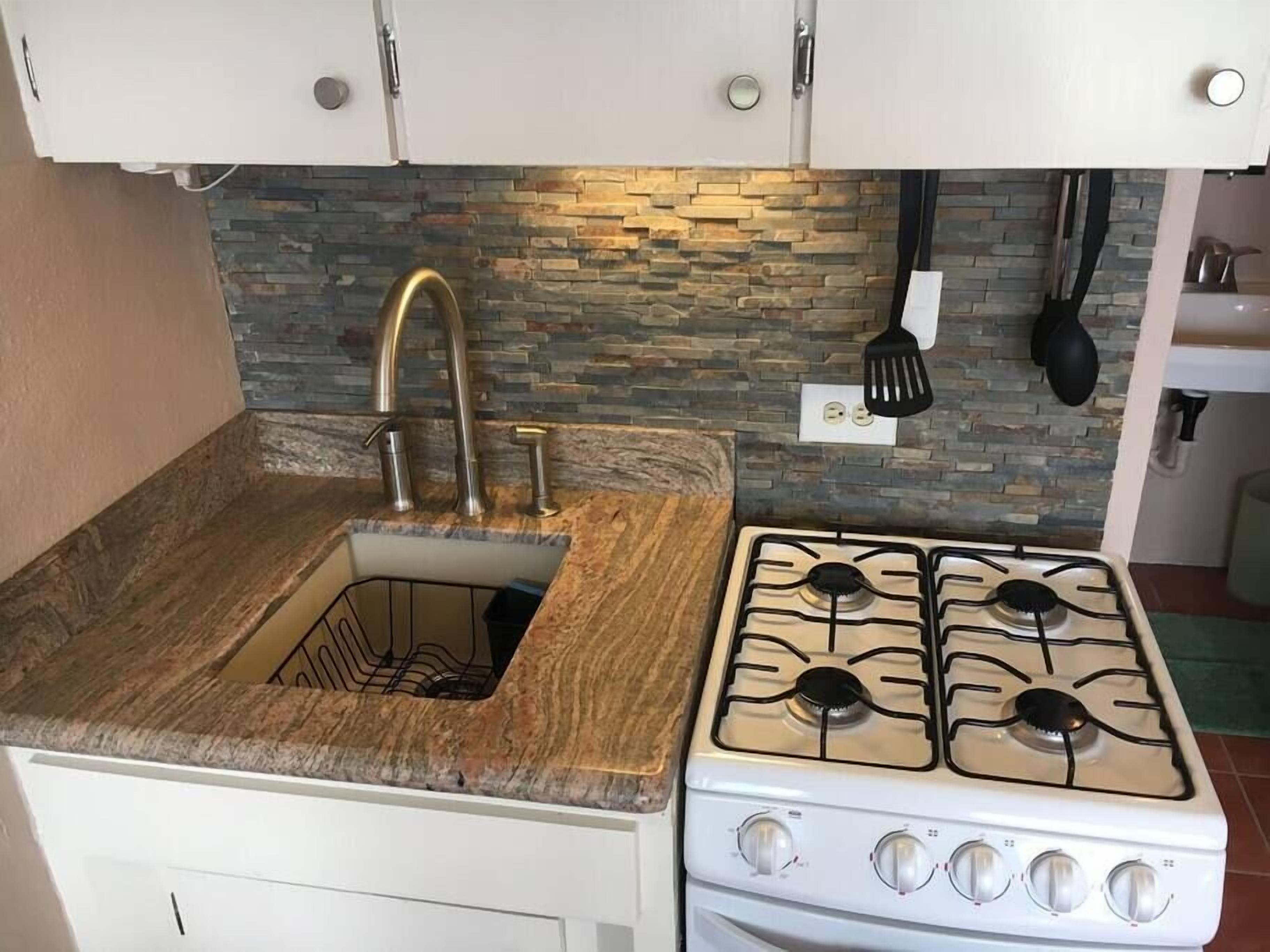 A kitchen with a granite countertop, a sink with a faucet, a stove, and a backsplash with a stone pattern.