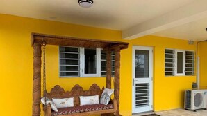 Um balanço de madeira com almofadas em frente a uma parede amarela, uma porta branca e um aparelho de ar condicionado.