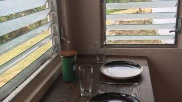 A table with two plates and a knife, a glass, and a green container by a window with blinds.