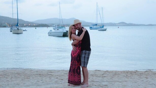Um casal se abraça em uma praia, com barcos ao fundo.