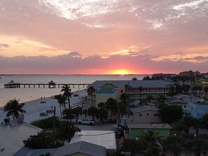A coastal town at sunset with a pier extending into the water, colorful buildings, and a beach with people and parked cars. - Escape to Cape! Heated Private Pool Home (Cape Coral)