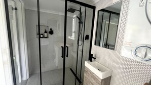Une salle de bain moderne dotée d’une douche à parois de verre, d’un lavabo blanc et d’un miroir avec un thermostat rond.