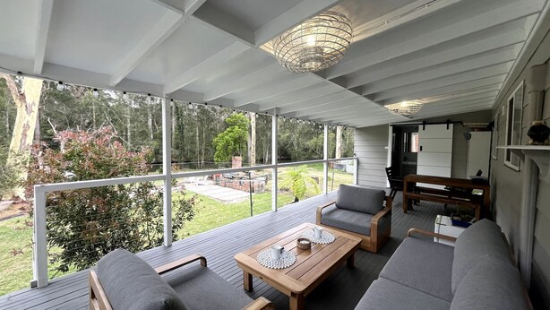 Un espace extérieur aménagé, abrité et doté d’une table en bois et de chaises rembourrées grises, offrant une vue sur un jardin et une structure en briques.