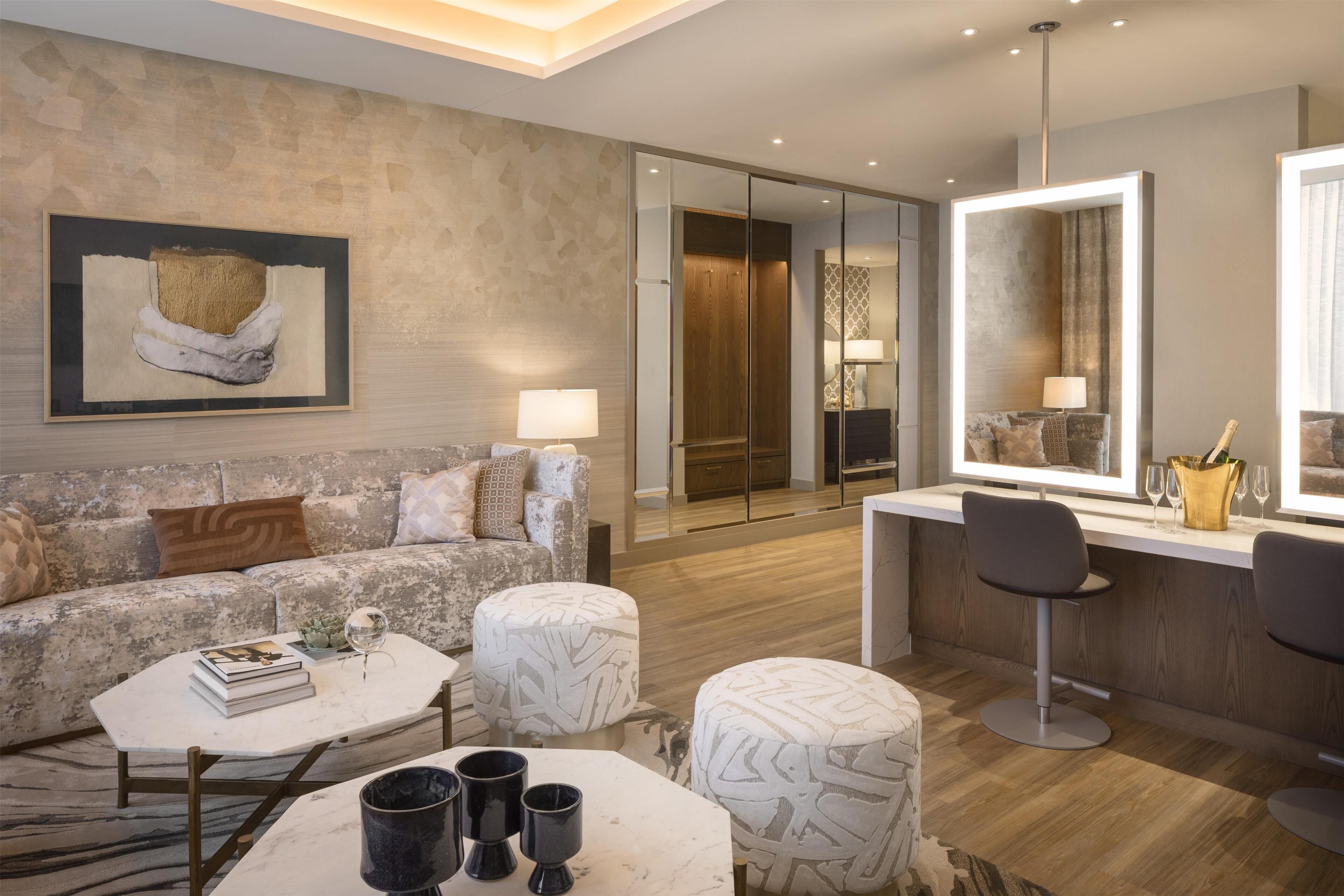 Una sala de estar moderna con sofá, mesa de centro y reposapiés, un escritorio con espejo, y una zona de bar con taburetes.