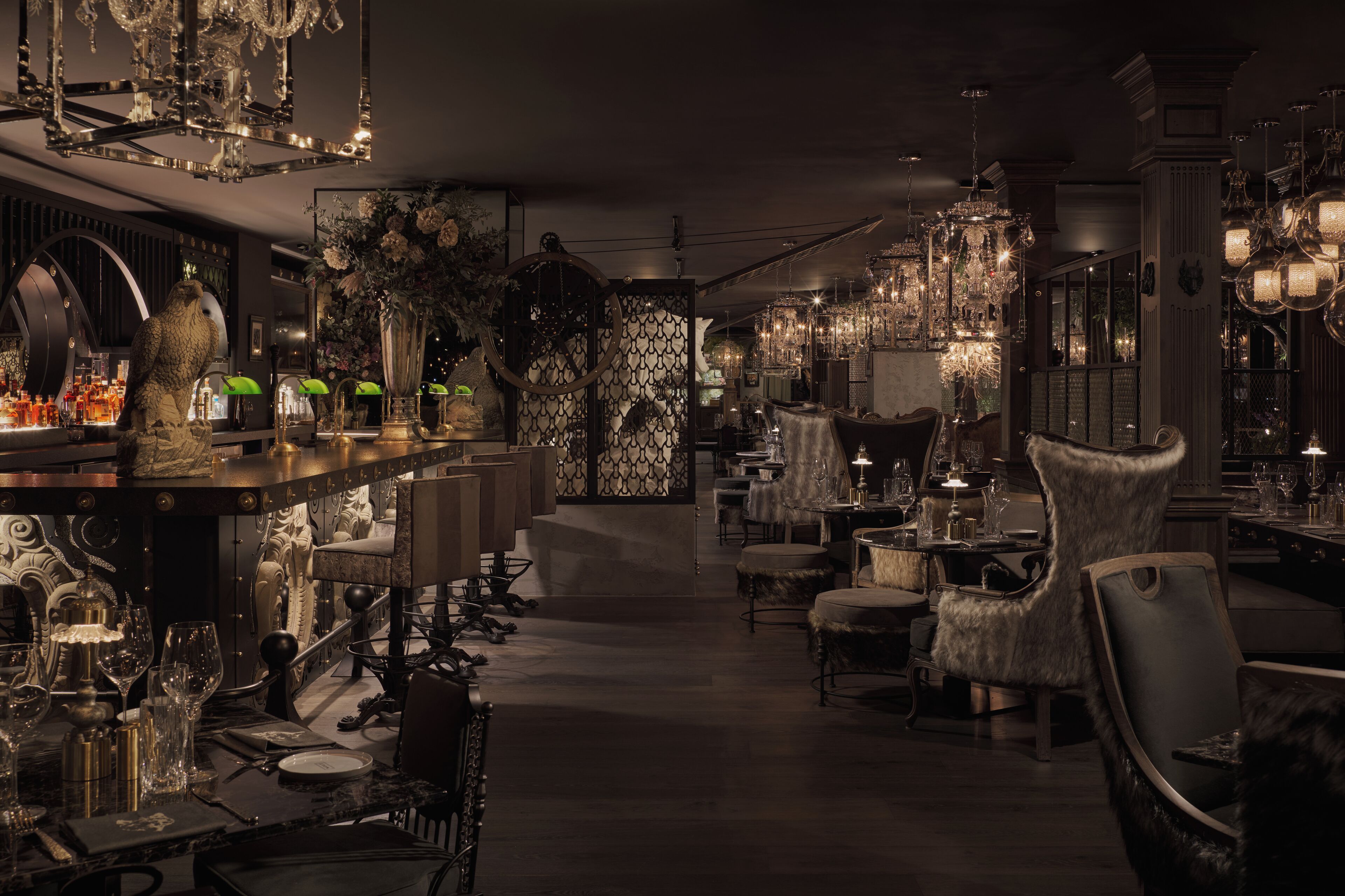 A dimly lit bar area with ornate chandeliers, plush seating, and a variety of bottles and glasses on display.