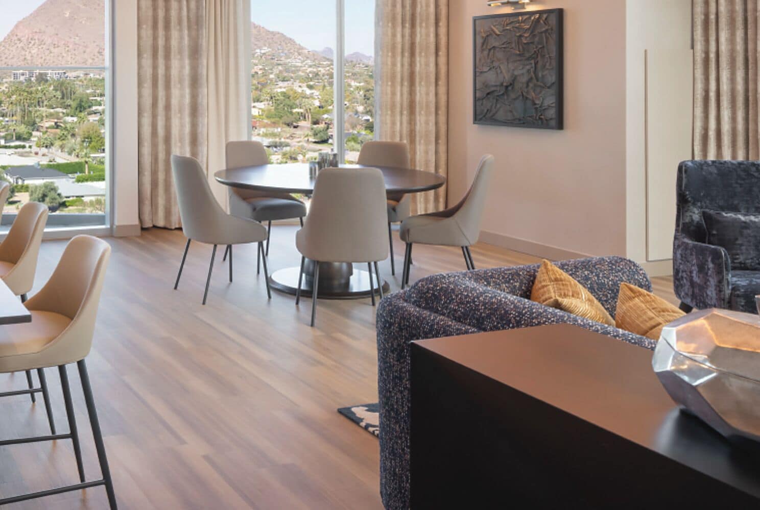 A modern hotel room with a dining area, a sofa, and a view of a mountainous landscape.