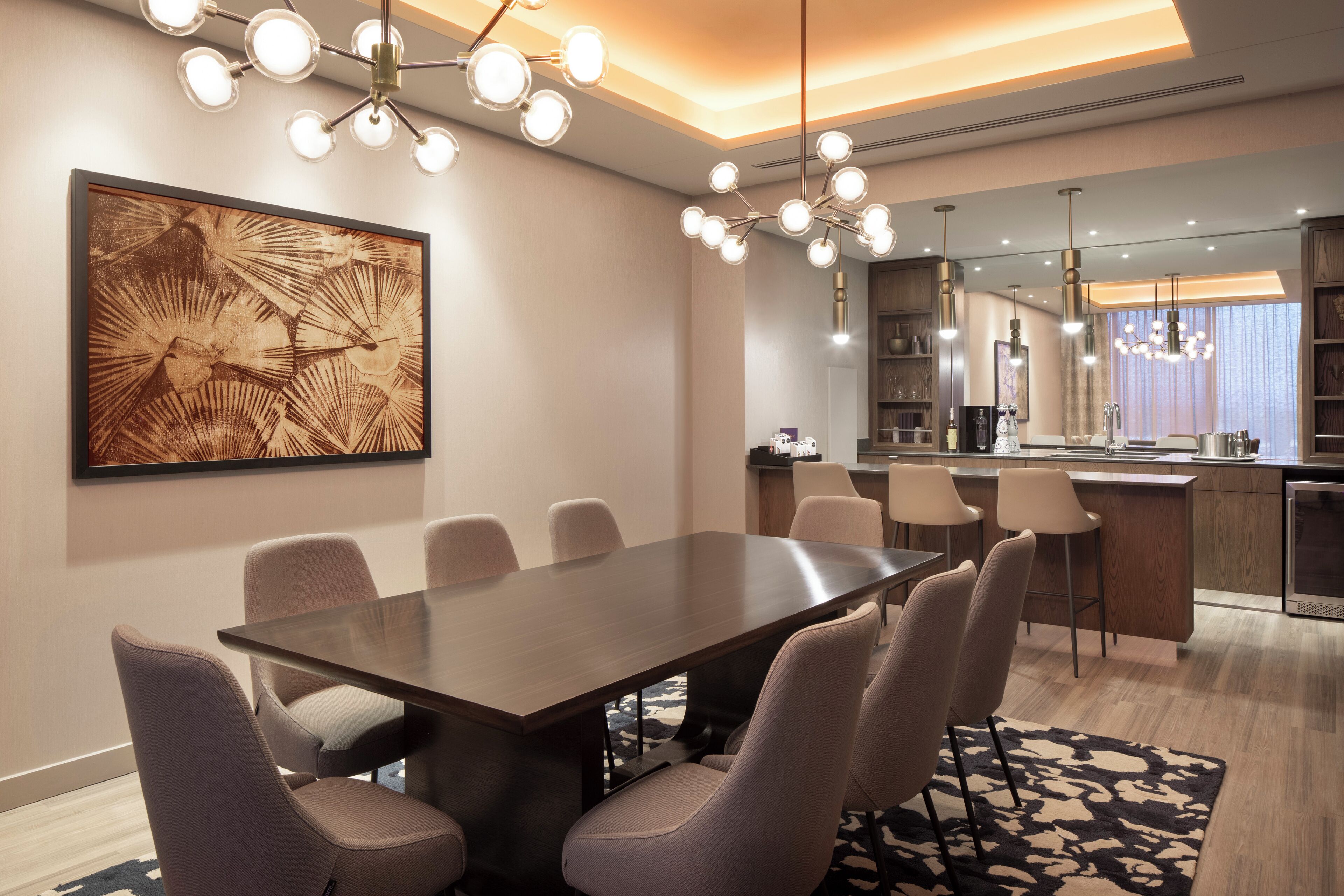 A modern dining area with a long table and chairs, a large abstract painting, and a kitchen in the background.