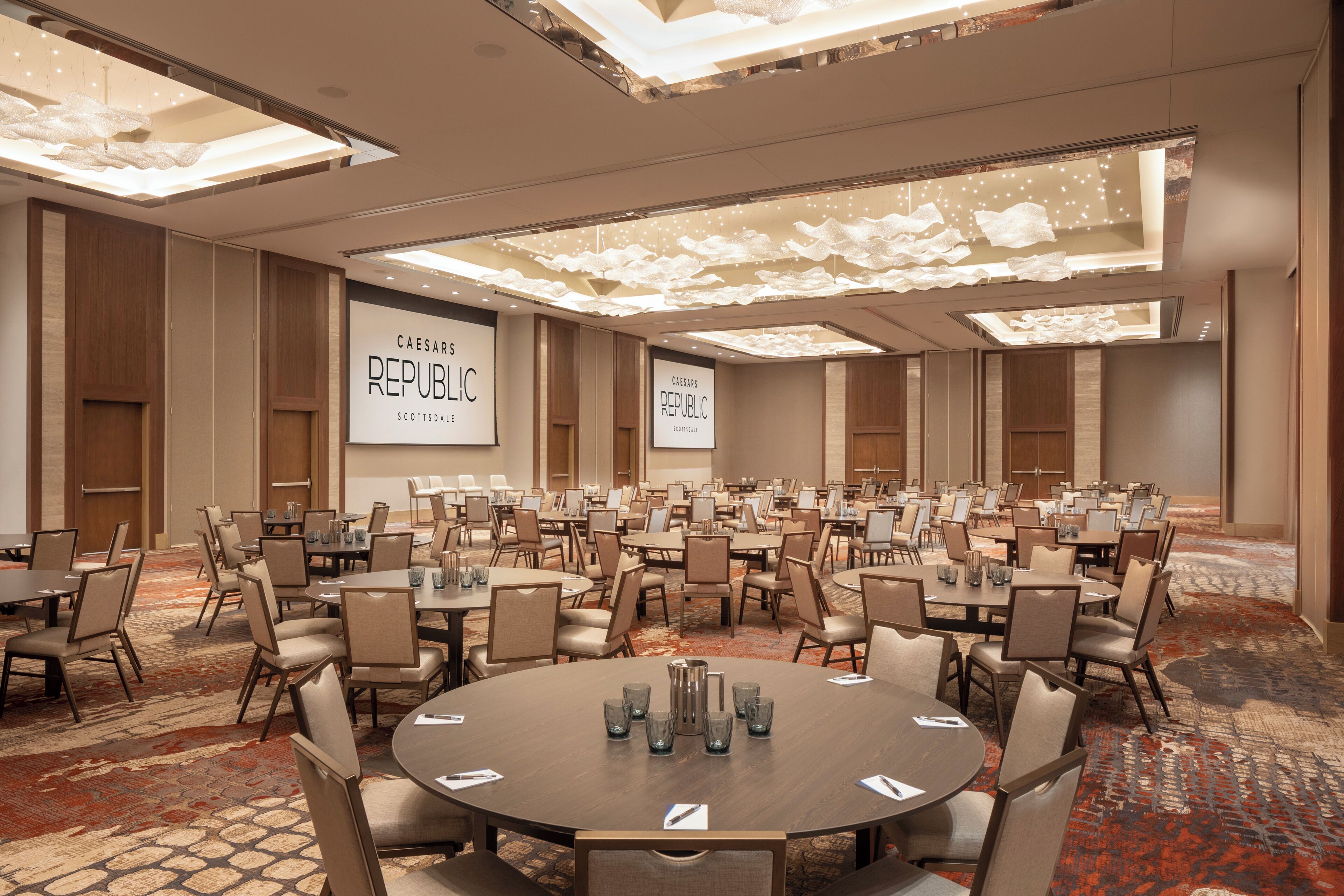 Un salón de conferencias con varias mesas y sillas, una pantalla grande mostrando 'Caesars Republic', y piso alfombrado.