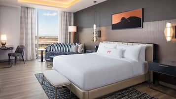 A modern hotel room with a large bed, a desk, a chair, a sofa, and a view of the cityscape.