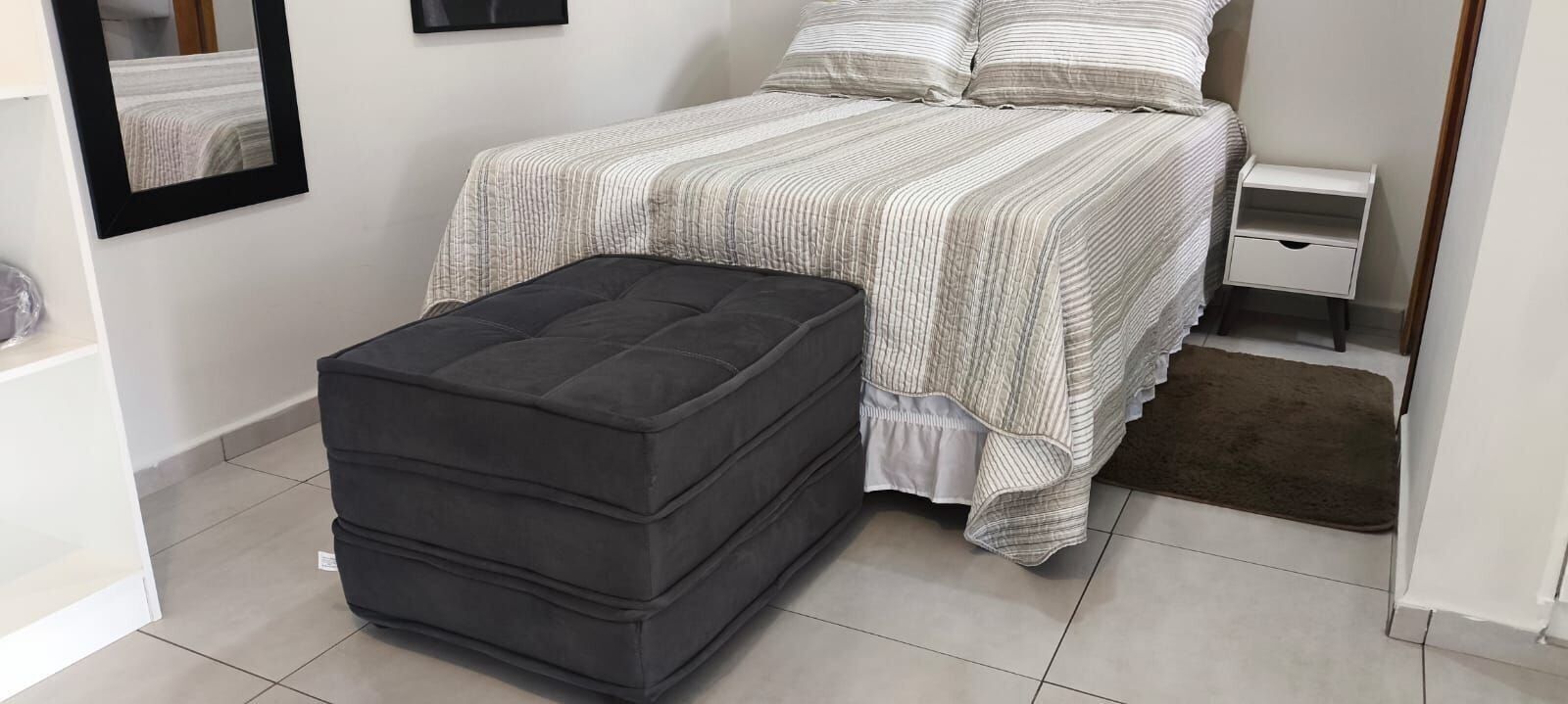 Een slaapkamer met een bed, nachtkastje en een grijze poef.