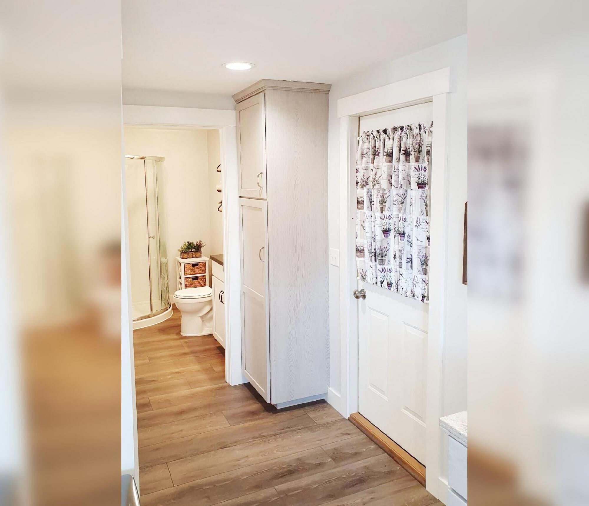 Un pasillo estrecho con suelo de madera, una puerta blanca con cortina de motivos vegetales y un armario blanco con un pequeño estante.