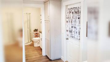 Un pasillo estrecho con suelo de madera, una puerta blanca con cortina de motivos vegetales y un armario blanco con un pequeño estante.