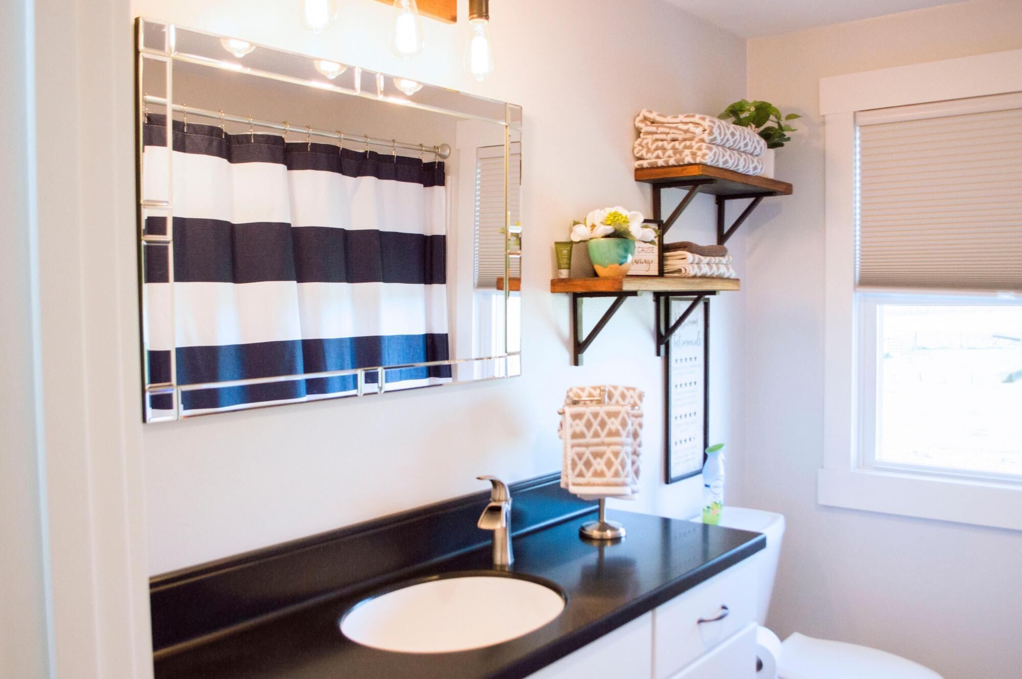A bathroom with a black countertop sink, a mirror with a striped shower curtain, and a shelf with towels and a potted plant.