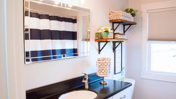 A bathroom with a black countertop sink, a mirror with a striped shower curtain, and a shelf with towels and a potted plant.