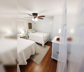 Une chambre avec deux lits, un ventilateur de plafond, une table de chevet avec une lampe et un petit meuble.
