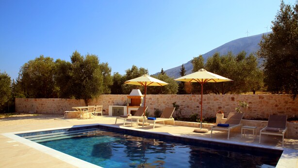 A swimming pool with lounge chairs and umbrellas, surrounded by stone walls and trees. - Unity Villa
 (KEFALONIA)