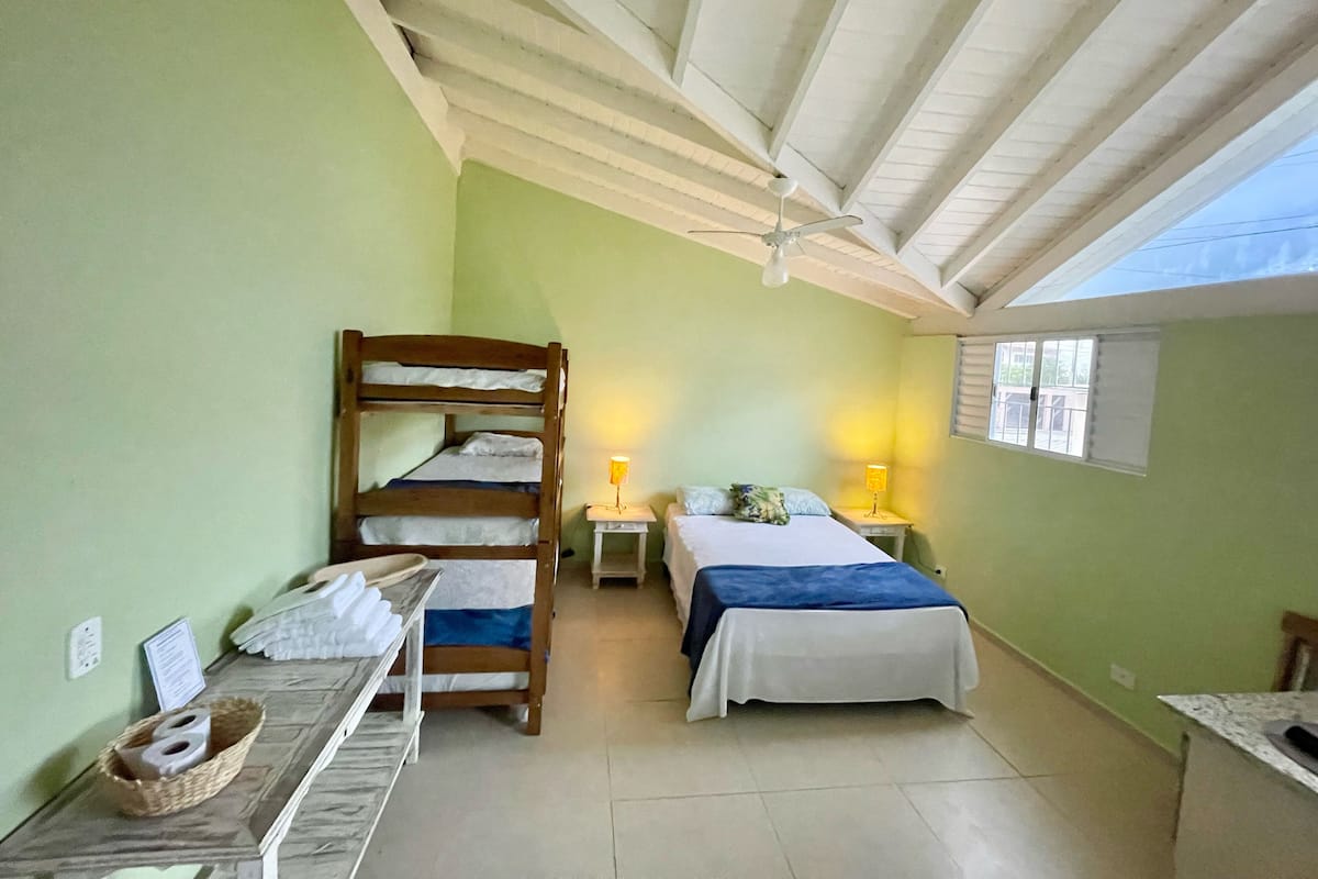 A small, cozy room with a bunk bed, a single bed, a desk with a lamp, and a window with blinds.