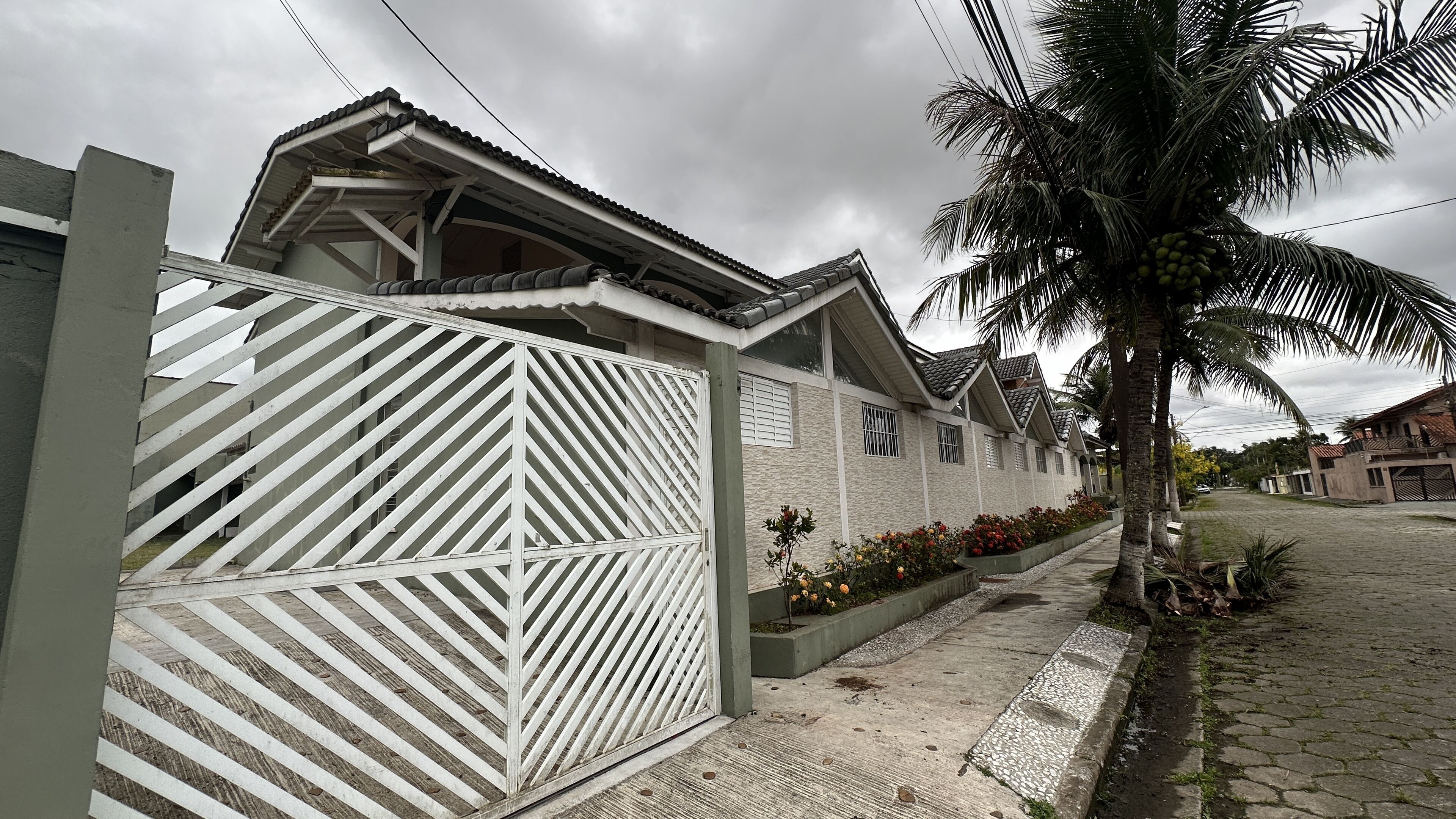 Edifício residencial com portão fechado, telhado cerâmico e caminho pavimentado com canteiros de flores.