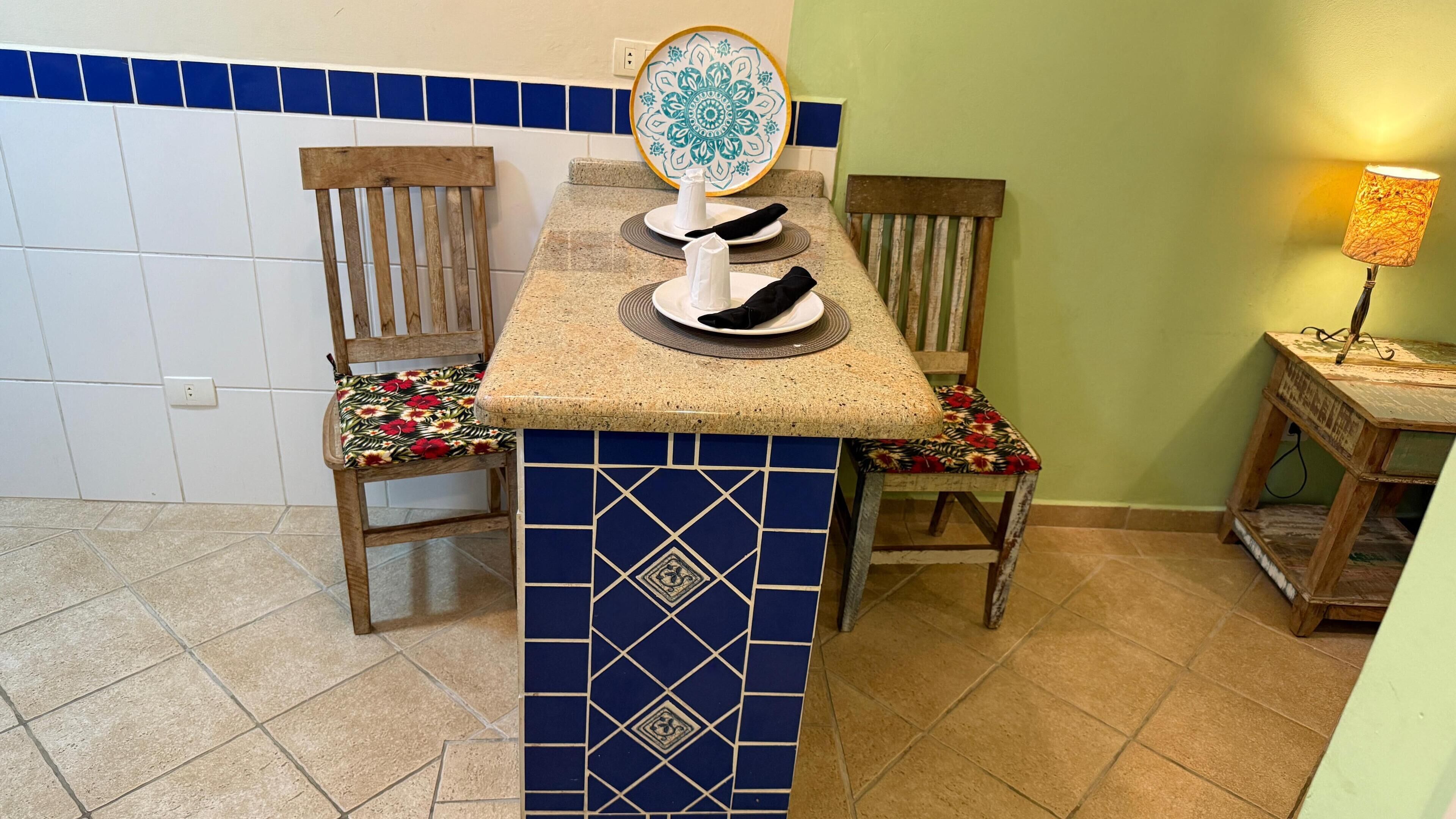 Une cuisine avec un mur recouvert de carreaux bleus et blancs, deux chaises en bois dotées de coussins à motifs floraux, un comptoir en granit et une assiette décorative accrochée au mur.