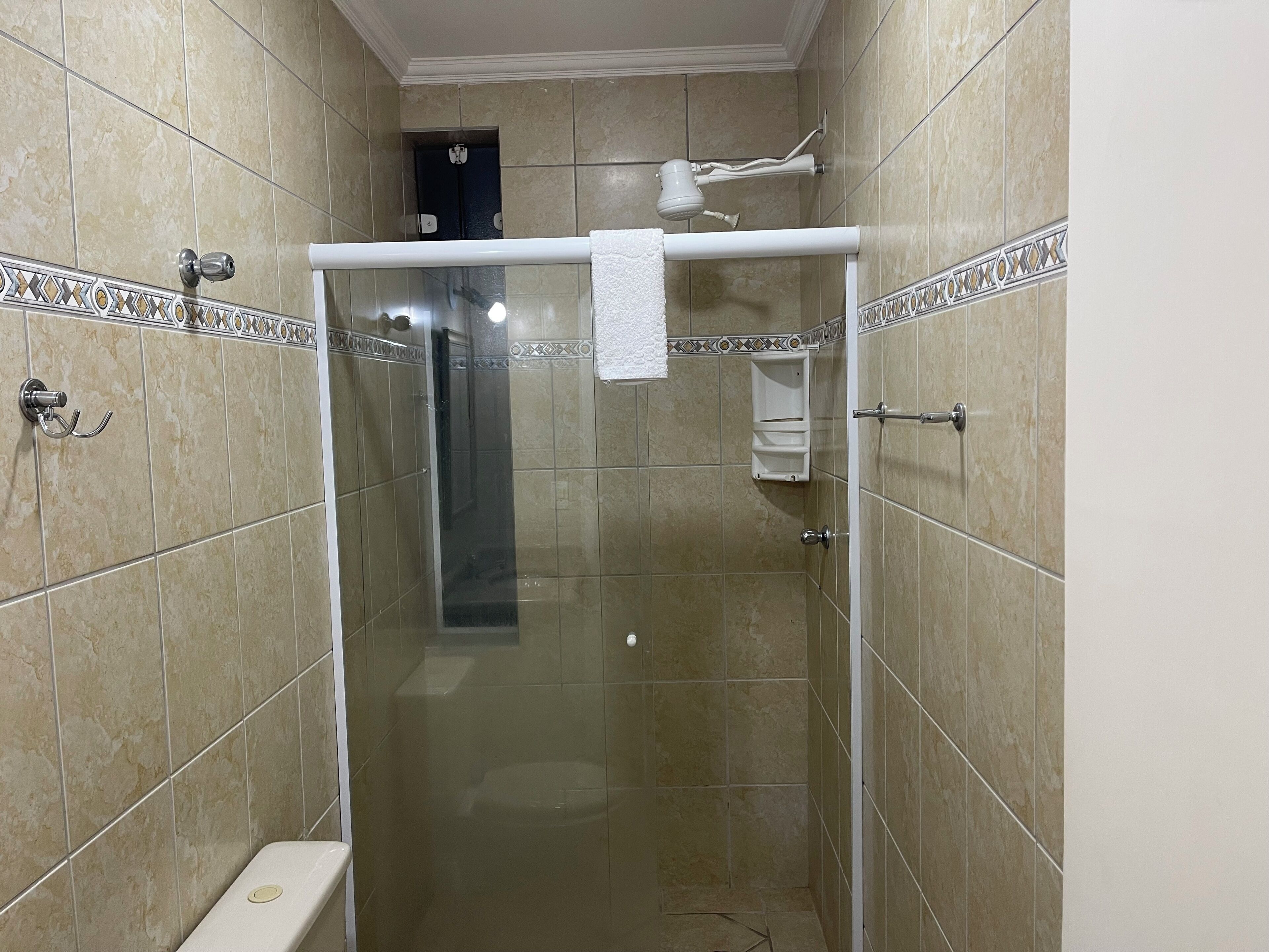 A glass-enclosed shower stall with a white towel hanging on a rack, beige tiled walls, and a visible toilet in the background.