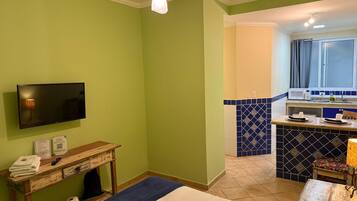 A room with a television, a wooden console table with a remote and towels, a blue rug, and a kitchen area with blue and white tiles.