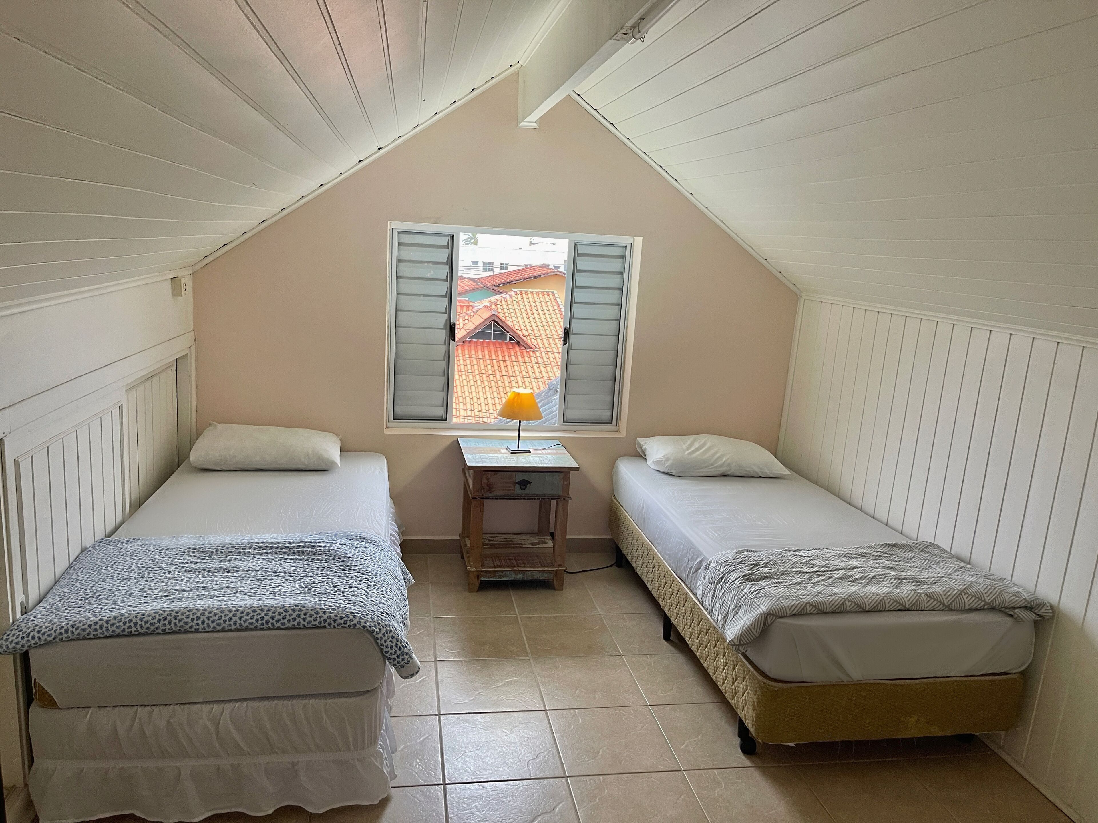 Una habitación pequeña y acogedora con dos camas individuales, una mesita con lámpara y una ventana con contraventanas.