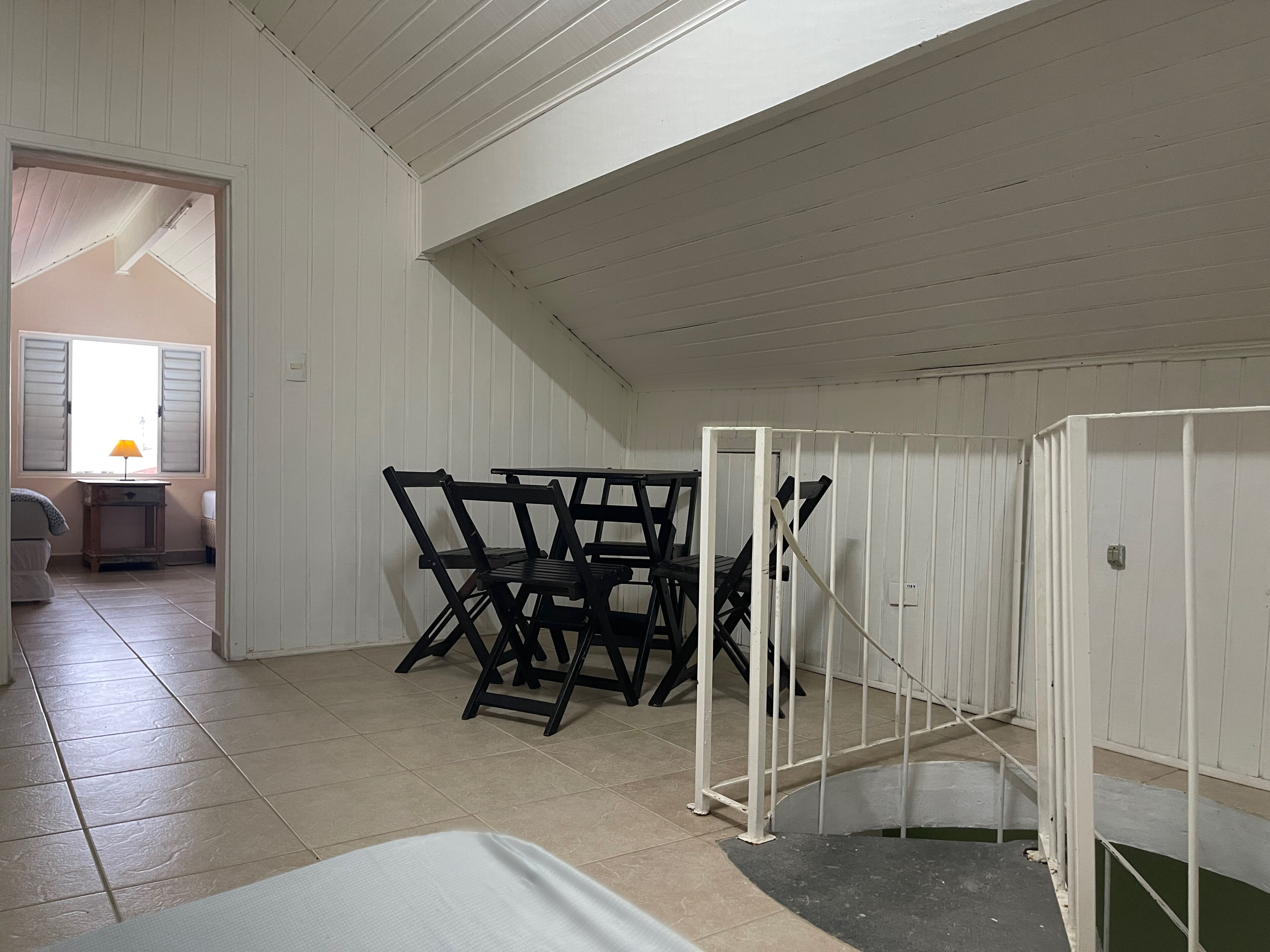 A room with a high ceiling, white walls, and a tiled floor. There is a staircase with a white railing leading to an upper level.