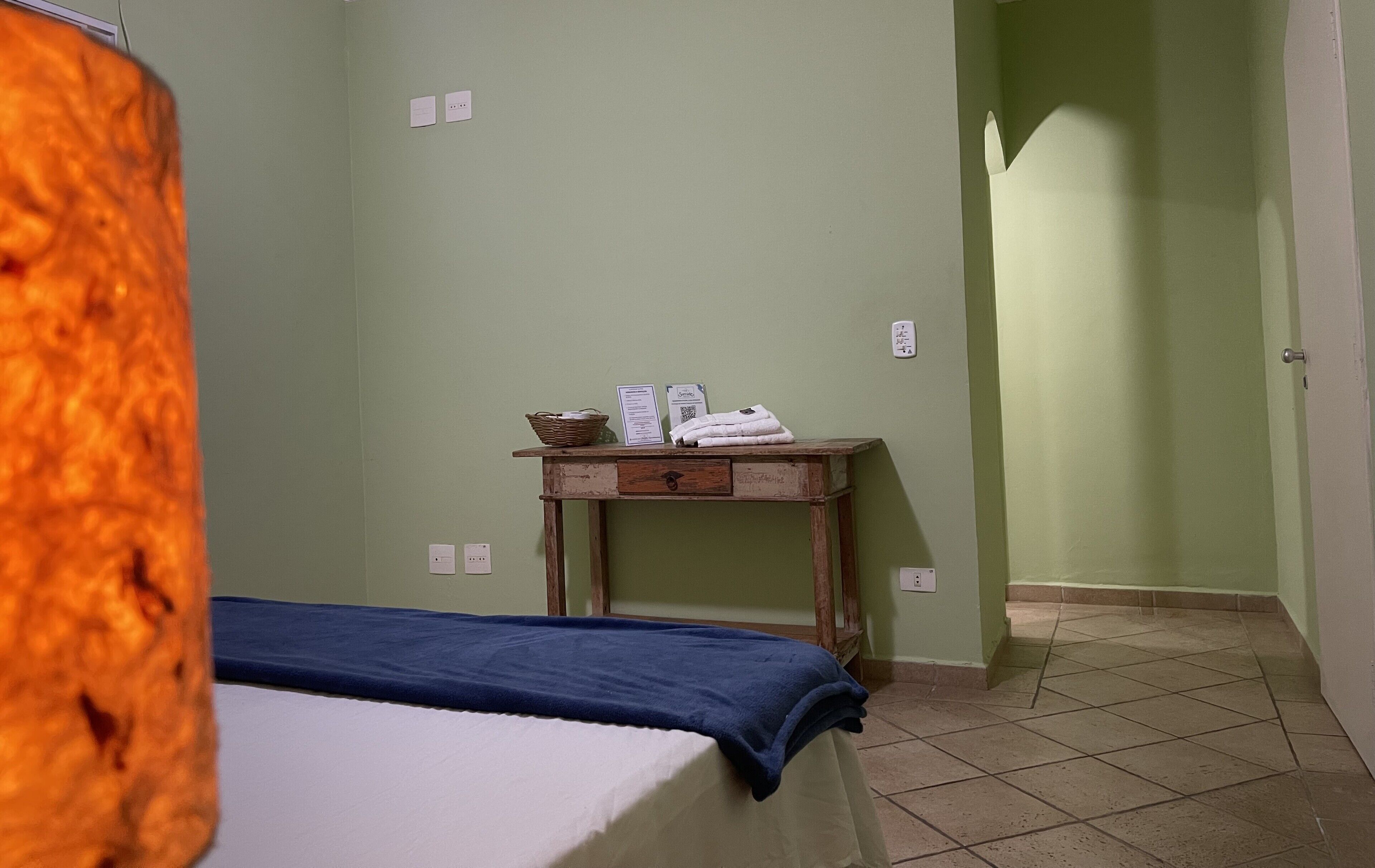 A room with a bed, a small wooden table with a basket and towels, and a door leading to another room.