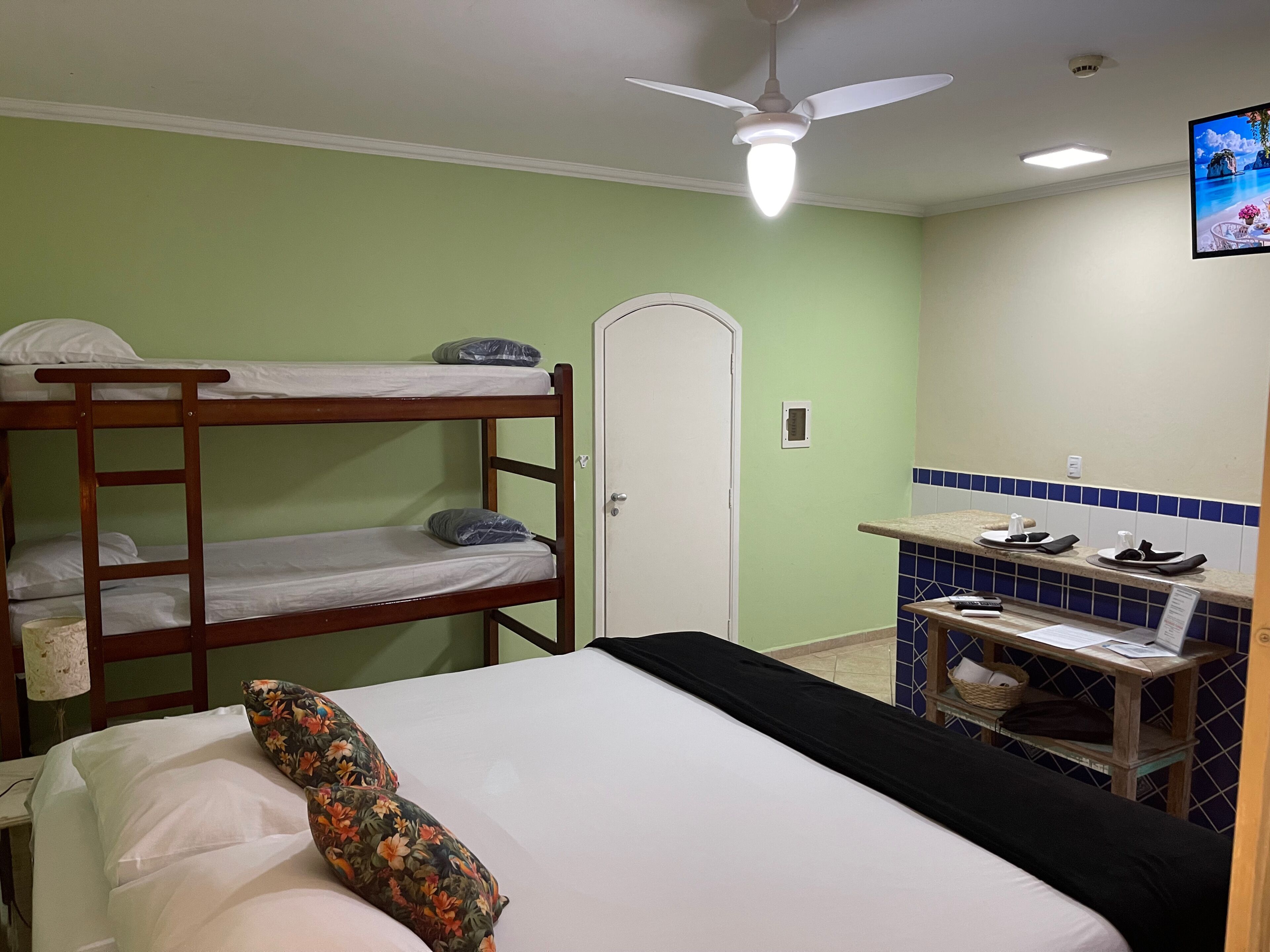 A bunk bed with a floral pillow, a single bed with a black bedspread, a ceiling fan, a small kitchenette with a tiled backsplash, and a door with a handle.