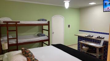 A bunk bed with a floral pillow, a single bed with a black bedspread, a ceiling fan, a small kitchenette with a tiled backsplash, and a door with a handle.