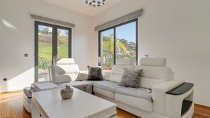 A modern living room with a white sectional sofa, a white coffee table, and a large window offering a view of a hillside. - Countryside Charm  (Camara de Lobos)