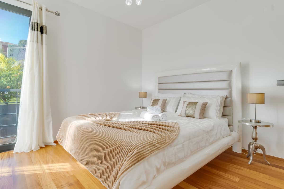 A modern bedroom with a large bed, a headboard, bedside tables, and a view of greenery outside.
