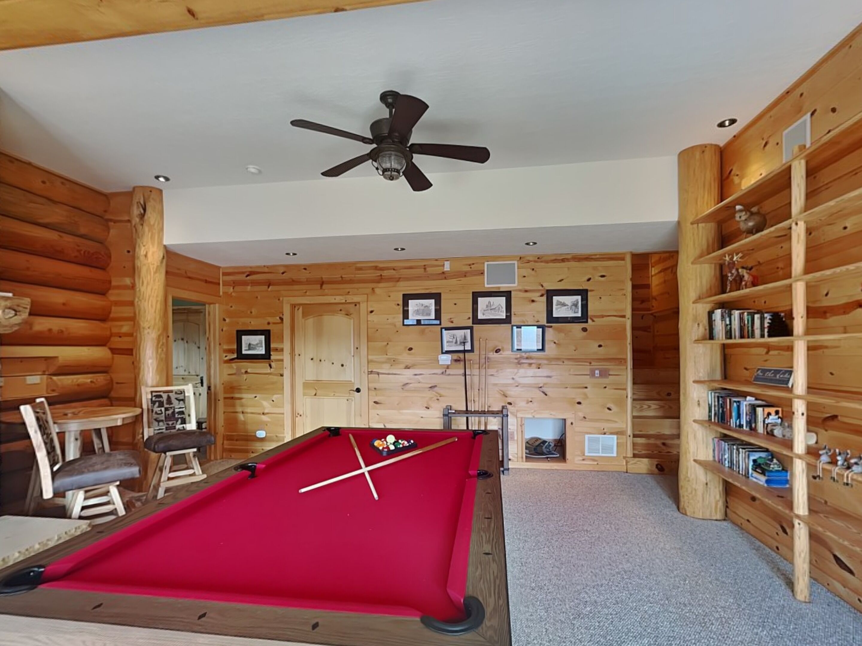 Une table de billard dans une pièce aux murs en bois, avec un ventilateur de plafond et des étagères à livres.