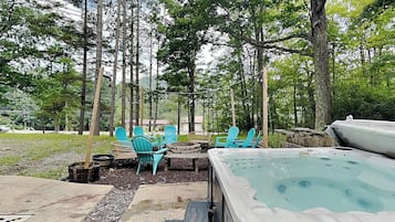 A hot tub surrounded by outdoor seating and a stone fire pit, set in a wooded area.