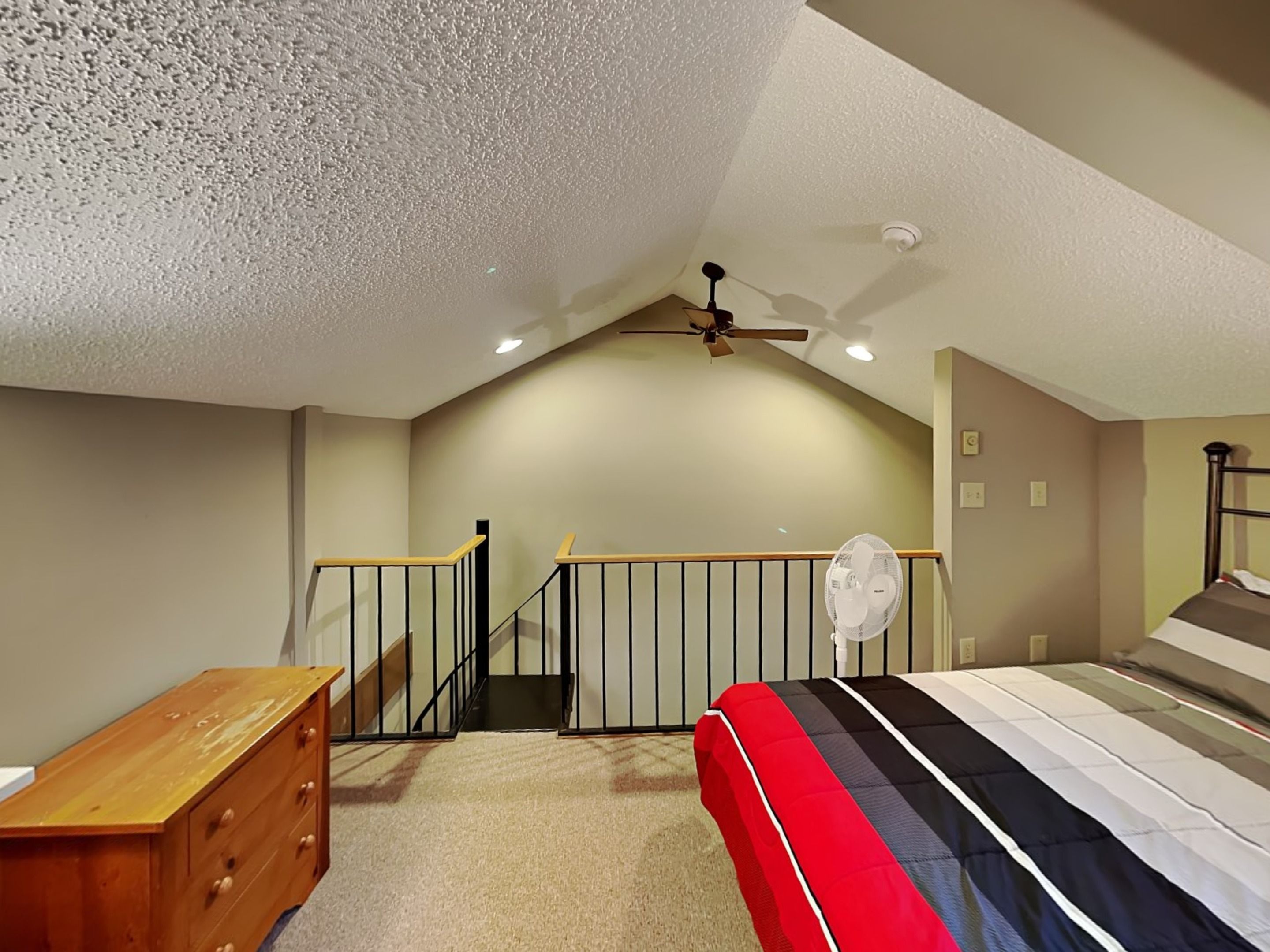 A bedroom with a sloped ceiling, a bed, a dresser, a fan, and a staircase.