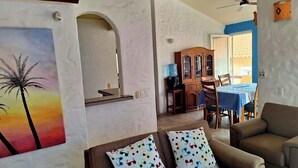 Una sala de estar con un sofá, un cuadro de palmeras, un comedor con una mesa y sillas, y un ventilador de techo.
