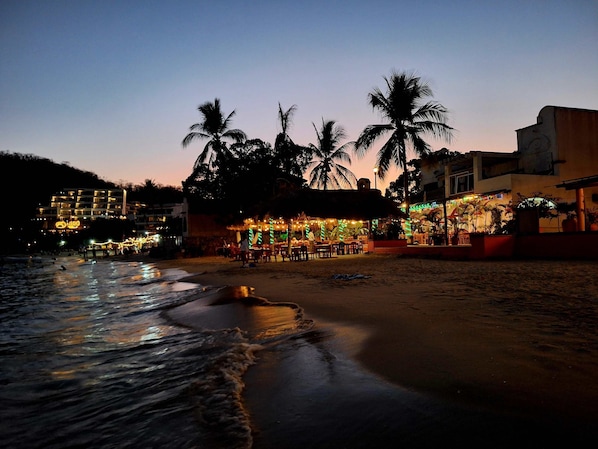 A beachfront hotel with a thatched-roof bar area, palm trees, and a view of the ocean.